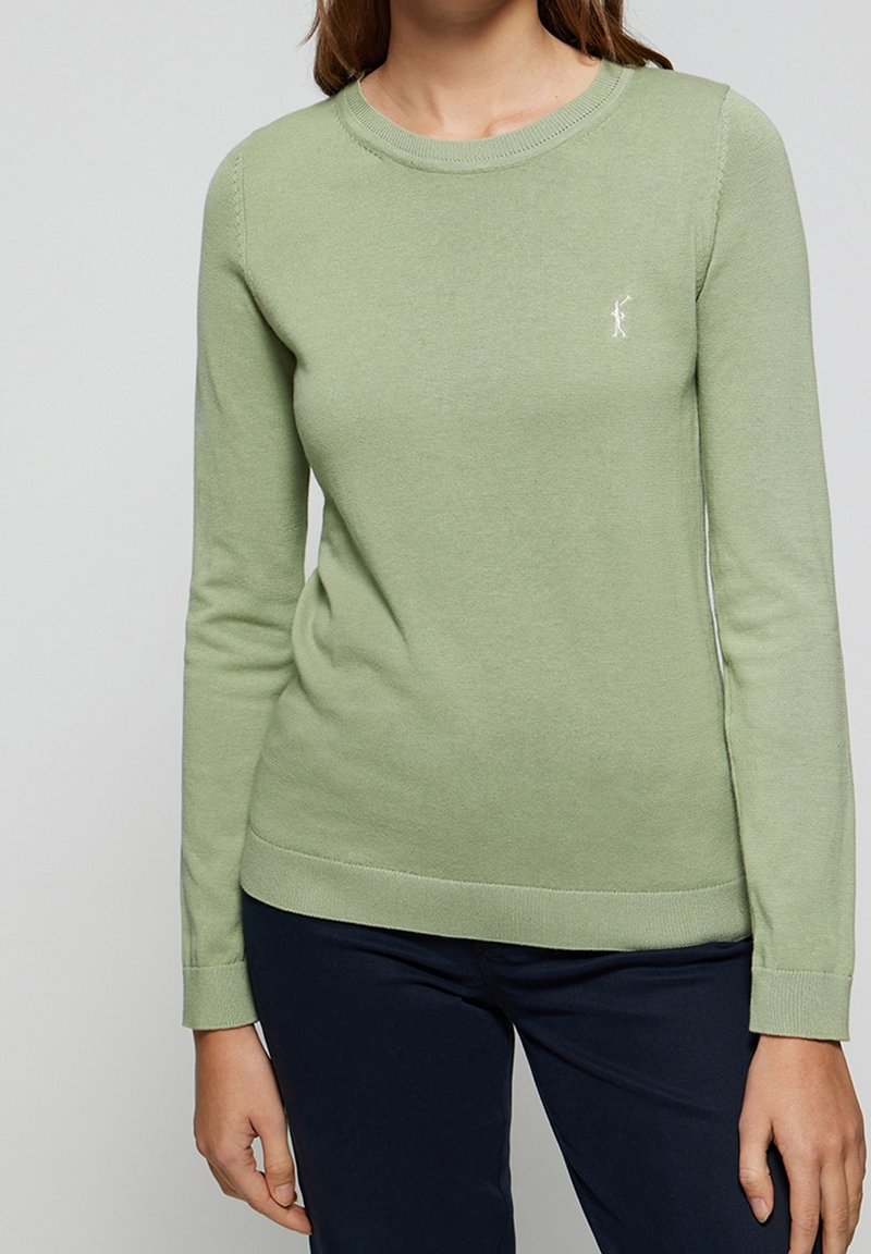 Femme portant un pull vert clair à manches longues avec un petit logo blanc sur la poitrine et un pantalon foncé, debout devant un fond uni.