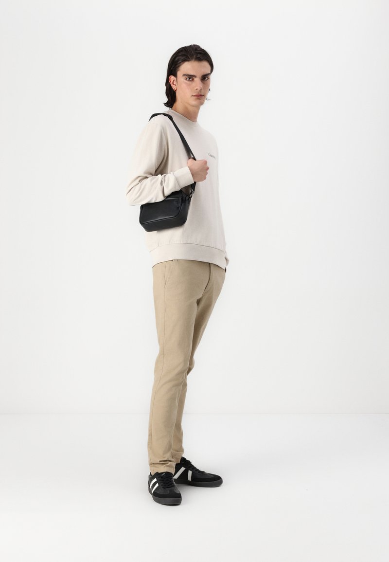 Sudadera beige claro con logo, pantalones beige, y zapatillas negras con rayas blancas; sosteniendo un pequeño bolso negro.