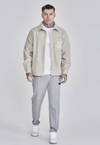 Light gray flannel shirt with a pocket logo, worn over a white T-shirt and paired with gray joggers and white sneakers. Neatly fitted design.