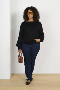 Pull en tricot noir avec des manches côtelées, jeans bleu foncé, ballerines noires et un petit sac à main marron. Cheveux bouclés et lunettes de soleil jaunes complètent le look.
