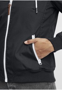 Black zip-up jacket featuring a white zipper, contrasting ribbed cuffs, and a pocket with a zipper. Smooth fabric with a durable finish.