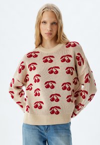 Beige gebreide trui met een rood kersenpatroon, ronde hals en geribbelde zoom. Het ontwerp heeft een comfortabele pasvorm met verlaagde schouders.