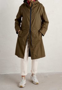 Olivgrön lättviktsjacka med huva, framdragkedja och sidofickor. Kombination med krämfärgade byxor och ljusa sneakers.