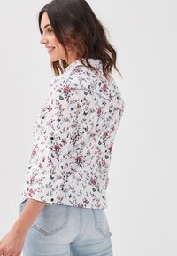 Blouse florale blanche avec des motifs rouges et bleus représentant des papillons, dotée d'un design boutonné et d'un col. Associée à un jean en denim bleu clair.