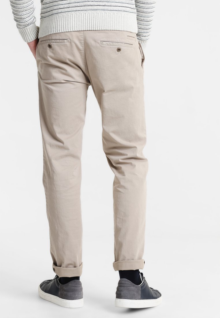 Beige Baumwollchinos mit schmalem Schnitt, ausgestattet mit Gesäßtaschen und umgeschlagenen Säumen, kombiniert mit grauen Sneakers auf einem weißen Hintergrund.