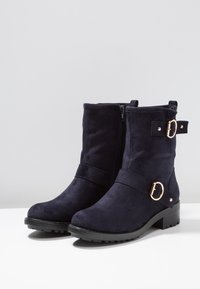 Botas de tobillo de ante azul marino con puntera redondeada, tacón bajo en bloque, cremallera lateral y adornos decorativos de hebilla en tono plateado.