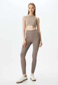 Lichtbruine cropped tanktop en bijpassende hoog-getailleerde legging, gemaakt van rekbare stof, gecombineerd met witte sneakers voor een sportieve uitstraling.