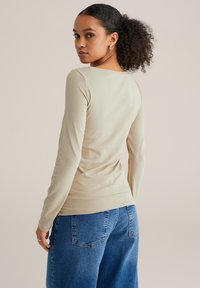 Smėlio spalvos ilgomis rankovėmis marškiniai su aptemptu siluetu, lygiu paviršiumi ir apvalia iškirpte, derinami su laisvomis mėlynomis džinsinėmis kelnėmis, kurios turi galines kišenes.