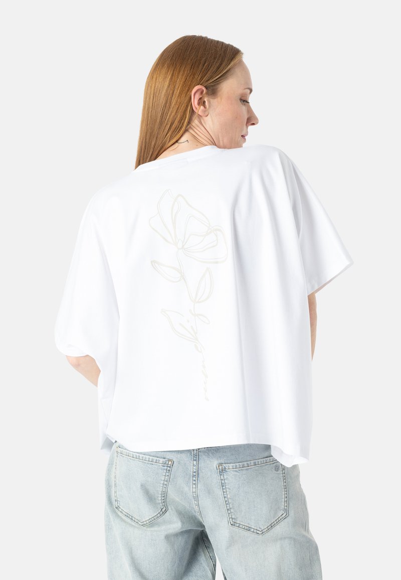Femme aux longs cheveux roux vue de profil, portant un t-shirt blanc ample avec un délicat motif floral en ligne à l'arrière et un jean bleu clair.