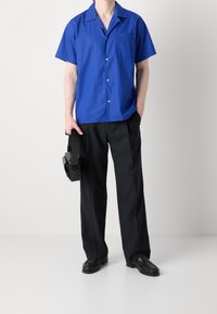 Niebieska koszula z krótkim rękawem i kieszenią naszytą, zestawiona z czarnymi, prostymi spodniami. Model trzyma czarne słuchawki w jednej ręce.
