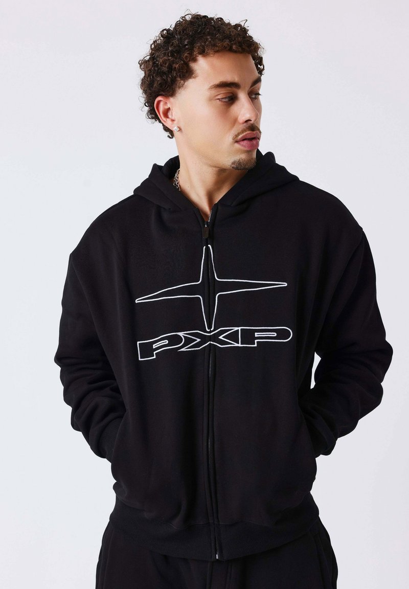 Homme aux cheveux bouclés regarde vers la droite, portant un sweat à capuche zippé noir avec un logo PXP blanc et les mains dans les poches, sur un fond uni.