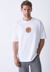 Hvid bomulds t-shirt med et rundt "Duff" logo i orange og sort, korte ærmer, afslappet pasform, parret med grå denim jeans.