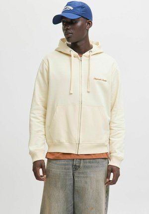 Sudadera cremosa con cremallera y cordones, y bolsillos frontales, con un pequeño logotipo bordado. Lleva puesta sobre una camisa naranja y combinada con jeans descoloridos.