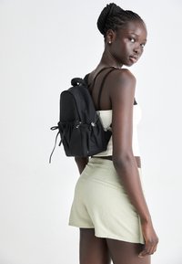 Black fabric backpack with cinched top, adjustable straps, and side hardware details. Worn on a model with braided hair and casual outfit.