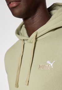 Vihreä puuvillapaita, jossa on kengurupocket, nyöripäällinen ja valkoinen Puma-logo brodeerattuna rinnassa.