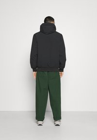 Veste noire avec capuche, poignets et ourlet élastiques. Associée à un pantalon large vert et des baskets de couleur claire. Vue de dos.