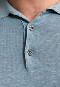 Světle modrá pletená polokošile s texturovaným povrchem, vybavená límečkem a dvěma kovovými knoflíky na léze.