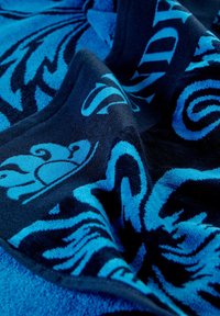 Toalla de playa azul con un diseño texturizado y patrones en azul oscuro contrastantes, exhibiendo motivos florales y abstractos. Fabricada con un tejido suave y absorbente.