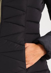 Zwarte donzen jas met chevron-gestikt patroon, voorzien van een gouden rits en een aansluitende kraag. De stof lijkt lichtgewicht en glad.