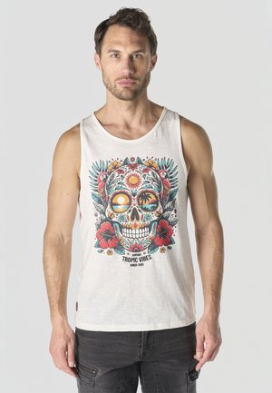 Hombre con camiseta sin mangas blanca con gráfico colorido de calavera tropical y loros, de pie contra un fondo claro y liso.