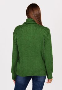 Vrouw met lang bruin haar, gekleed in een chunky groene coltrui en donkerblauwe jeans, staand met haar rug naar de camera tegen een witte achtergrond.