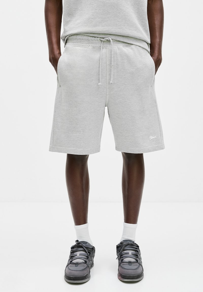 Mann trägt hellgraue Shorts mit Kordelzug, weiße Sportsocken und schwarze Turnschuhe, steht mit den Händen in den Taschen.