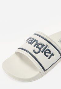 Sandale albe cu talpă plată, având o curea largă de culoare navy cu logo-ul "Wrangler", suprafață texturată și interior neted, concepute pentru confort.