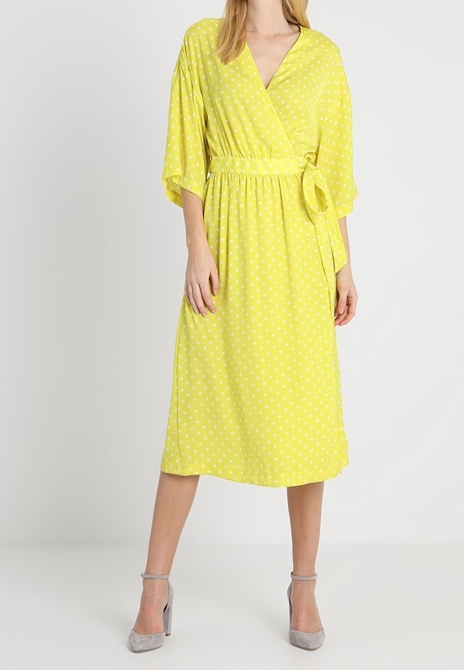 Femme portant une robe portefeuille jaune à pois blancs, manches courtes, taille nouée, et des escarpins gris, debout devant un fond uni.
