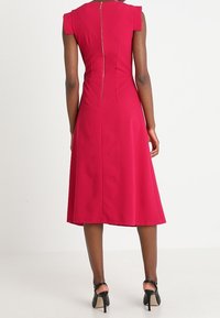 Robe rouge jusqu'aux genoux avec des manches courtes, un corsage ajusté et une fermeture éclair apparente au dos. Tissu lisse et jupe évasée.