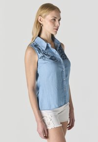 Mujer con camisa sin mangas de color azul claro con botones, detalles florales oscuros bordados y pantalones cortos blancos con ribete de encaje, de pie de lado.