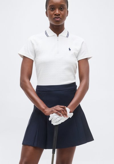 Γυναίκα με λευκό πόλο με φερμουάρ, μπλε φούστα με πιέτες και λευκά γάντια γκολφ κρατάει κάθετα μπαστούνι του γκολφ μπροστά της.