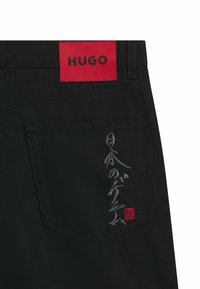 Schwarze Denim-Hosen mit einem roten Etikett, das 'HUGO' mit gesticktem grauen Text trägt, sowie einem kleinen roten Logo auf der Gesäßtasche.