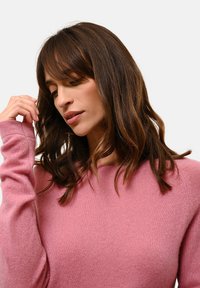 Růžový svetr vyrobený z jemného materiálu, s kulatým výstřihem a dlouhými rukávy s mírně priléhavými manžetami. Jemná textura je viditelná.