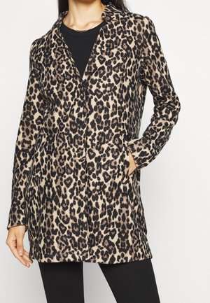 Femme portant un manteau mi-long à motif léopard avec col, poches, et un haut noir en dessous, se tenant devant un fond blanc.