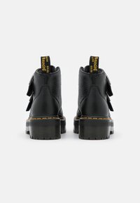 Bottines noires en cuir avec une finition texturée, semelle épaisse et coutures jaunes. Elles possèdent des tirants distincts et des sangles latérales ajustables.