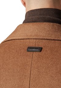 Brązowy płaszcz wełniany z miękką fakturą, z brązowym ściągaczem u kołnierza oraz czarną skórzaną metką marki na plecach.