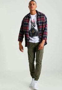 Geruite flanellen overhemd in rood en marineblauw over een witte T-shirt met opdruk. Olijfkleurige cargobroek met elastieken boorden en witte sneakers.