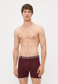 Boxers en bordeaux fabriqués en tissu extensible avec une taille noire présentant le texte "PIER ONE" en blanc. Texture lisse, design ajusté.
