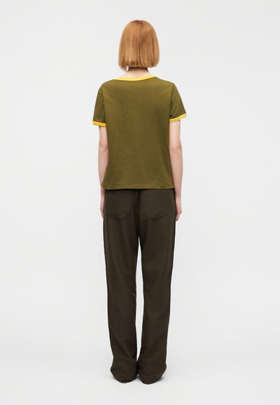 Φόρεμα χρώματος ελιάς με κίτρινο περίγραμμα, κοντά μανίκια και άνετη εφαρμογή, συνδυασμένο με χαλαρά καστανά σκούρα παντελόνια με πίσω τσέπες.