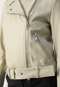 Jachetă de motocicletă din piele crem, cu un design ajustat, fermoare argintii și talie cu curea și închidere cu capse. Textură netedă, fără modele vizibile.