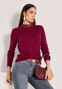 Frau in einem burgunderfarbenen Pullover mit verziertem Kragen, grauen Jeans, passendem Gürtel, die eine mit Nieten verzierte burgunderfarbene Handtasche hält und auffällige Ohrringe trägt.