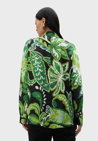 Svart blus med lång ärm i tyg som har stora gröna blommönster och blad. Inkluderar knäppning vid ärmslut och en hög krage.