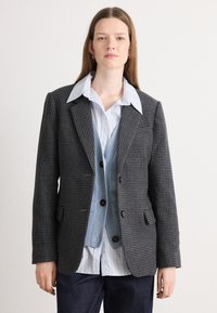 Blazer de cuadros grises con dos botones, usado sobre un chaleco de punto azul claro y una camisa blanca de rayas. Presenta un cuello clásico y bolsillos laterales.