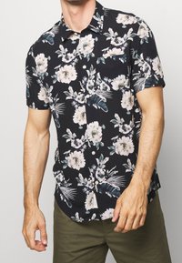 Camisa de flores de manga corta con fondo negro, que presenta flores blancas y verdes, hojas de palma y un diseño de botones en la parte delantera. Tejido texturizado.