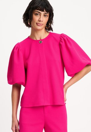 Vrouw met kort donker haar, gekleed in een felroze textuur top met pofmouwen en bijpassende broek tegen een effen witte achtergrond.