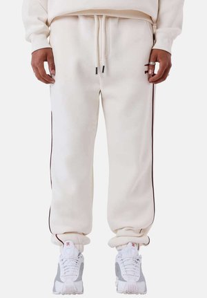 Personne portant un pantalon de survêtement écru avec des bandes noires sur les côtés, une taille à cordon et des baskets blanches-grises, debout les mains détendues le long du corps.