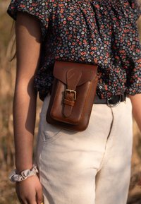 Bruine leren tas met een klep en gespdetails, bevestigd aan een beige corduroy tailleband, met een zwarte blouse met bloemenpatroon.