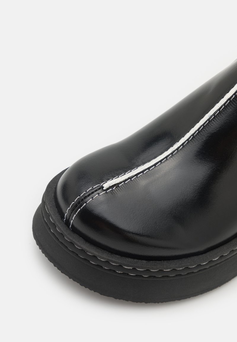 Bota de cuero negro con puntera redondeada, que presenta costuras blancas a lo largo de la costura y una gruesa suela de goma negra. Textura suave y brillante.