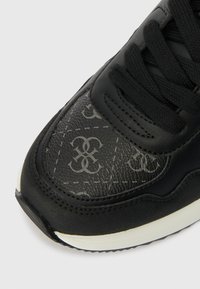 Melni sporta apavi ar teksturētu virsmu, kontrastējošu pelēku logotipa rakstu, auklu aizdari un baltu gumijas zoli.