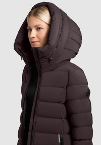 Puszysta zimowa kurtka w ciemnym śliwowym kolorze z dużym kapturem, z zamkiem błyskawicznym z przodu, teksturowanym materiałem oraz akcentem logo na rękawie.